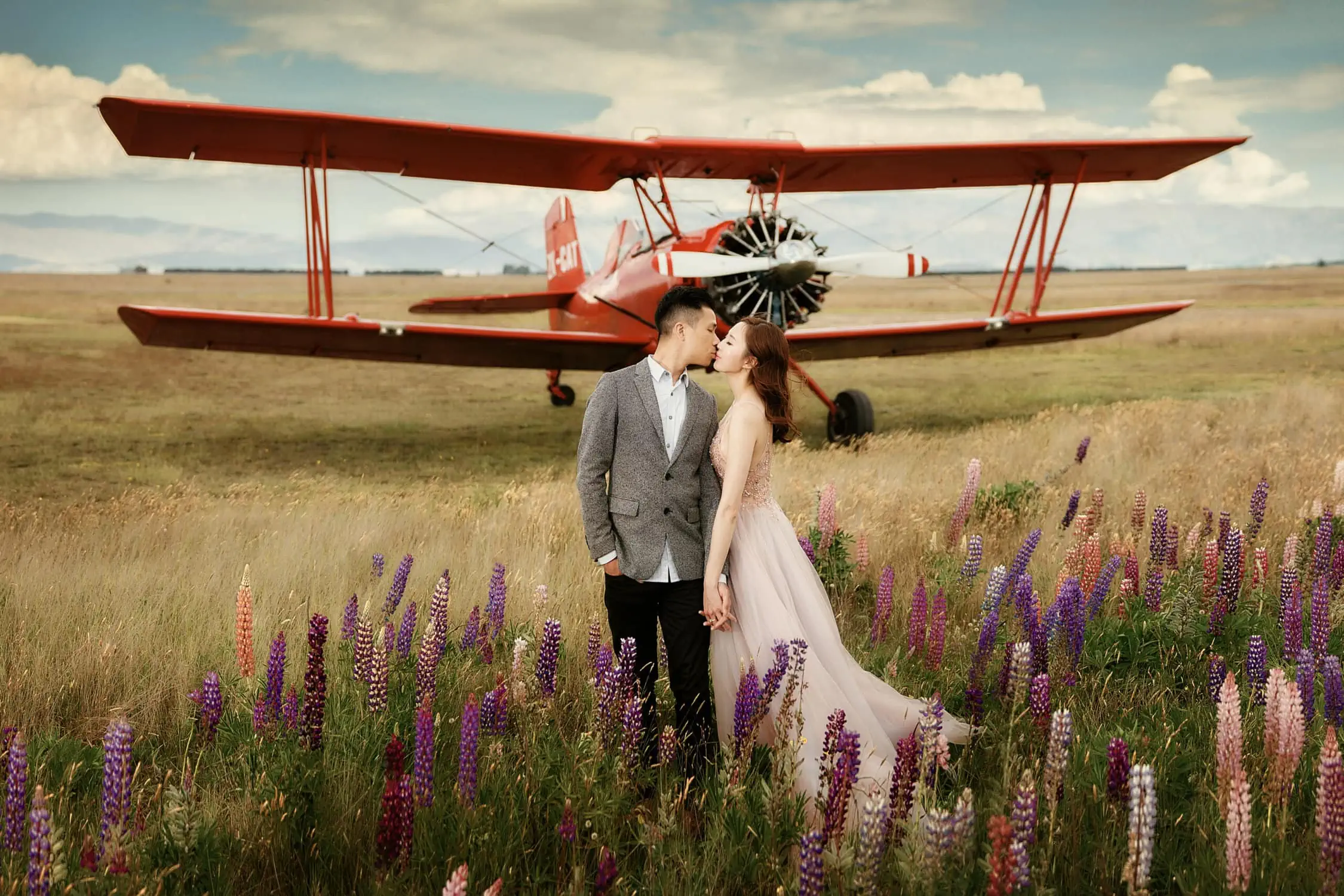 Queenstown Tekapo New Zealand Heli Wedding Elopement Pre-Wedding Shoot Photographer クイーンズタウン　テカポ　ニュージーランド　エロープメント 前撮り　フォトウェディング　結婚式 | A couple shares a kiss in a field of flowers with a vintage biplane in the background during their Tekapo pre-wedding shoot.